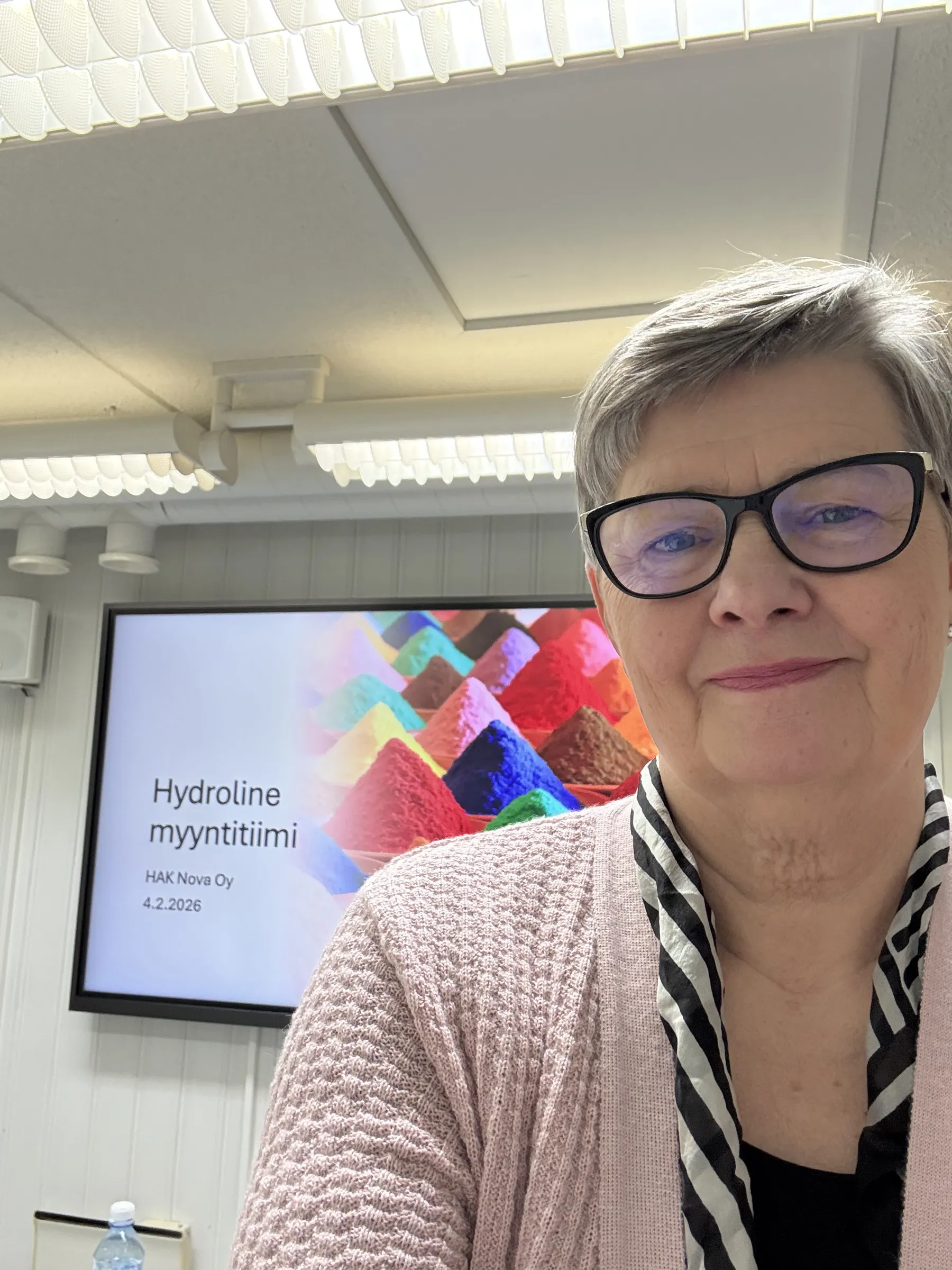 Lähikuva Hilkka Alatalo-Korvesta hymyilemässä. Hänellä on vaaleanpunainen neuletakki ja mustavalkoinen huivi. Taustalla näkyy kokoushuoneen valkoinen katto ja seinä, sekä näyttö, jossa on esityskalvo tekstillä 'Hydroline myyntitiimi' ja kuva värikkäistä jauhekasoista.
