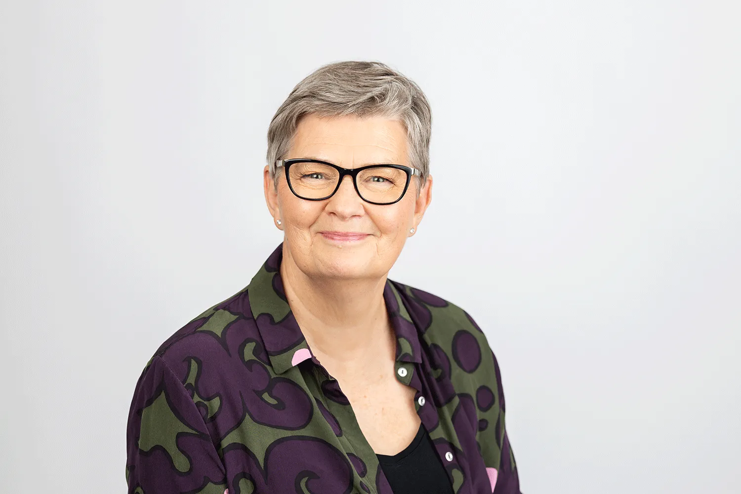 Lähikuva Hilkka Alatalo-Korvesta. Hänellä on yllään oliivinvihreä ja tummanvioletti kuviollinen paita, jonka alta pilkottaa musta toppi. Kuva on otettu tasaisessa studiovalossa vaaleaa taustaa vasten.